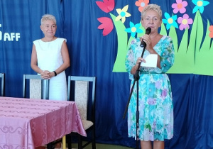 Barbara Siemienkowicz i Ewa Madalińska rozpoczynają uroczystość. Pani dyrektor stoi przy mikrofonie na tle dekoracji z kolorowych motyli i kwiatów.